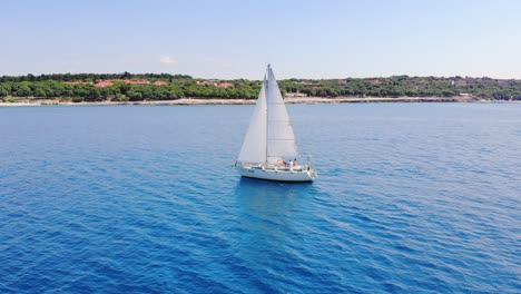 Lonely-white-sailboat-sails-along-green-coast-under-cloudless-skies-on-calm-clear-blue-water