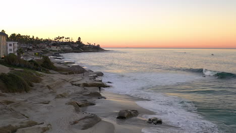 Surfer-with-surfboard-walks-on-beach-in-La-Jolla-at-sunrise,-drone-shot