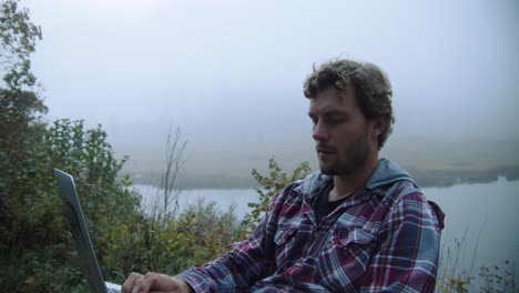 medium shot, young man using a laptop while looking around, foggy lake view in the background