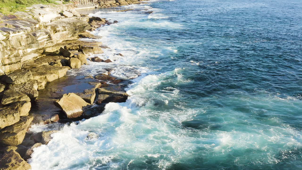 mientras las olas con flecos chocan contra las rocas del océano, el dron pov se mueve lentamente hacia atrás a lo largo de la cara del acantilado, la piscina del océano en el fondo, la playa de coogee, sydney, australia
