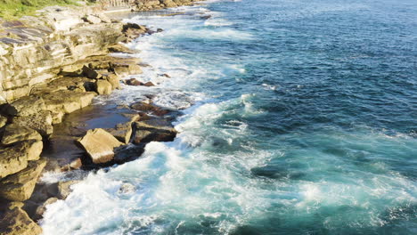 Mientras-Las-Olas-Con-Flecos-Chocan-Contra-Las-Rocas-Del-Océano,-El-Dron-Pov-Se-Mueve-Lentamente-Hacia-Atrás-A-Lo-Largo-De-La-Cara-Del-Acantilado,-La-Piscina-Del-Océano-En-El-Fondo,-La-Playa-De-Coogee,-Sydney,-Australia