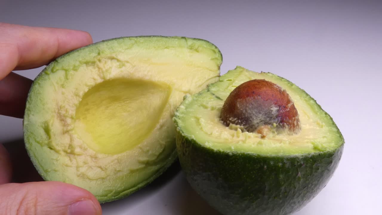 Um abacate peruano verde maduro sobre um fundo branco. Uma mão entra em cena e abre a fruta pré-fatiada. O caroço do abacate é de cor marrom-alaranjada.