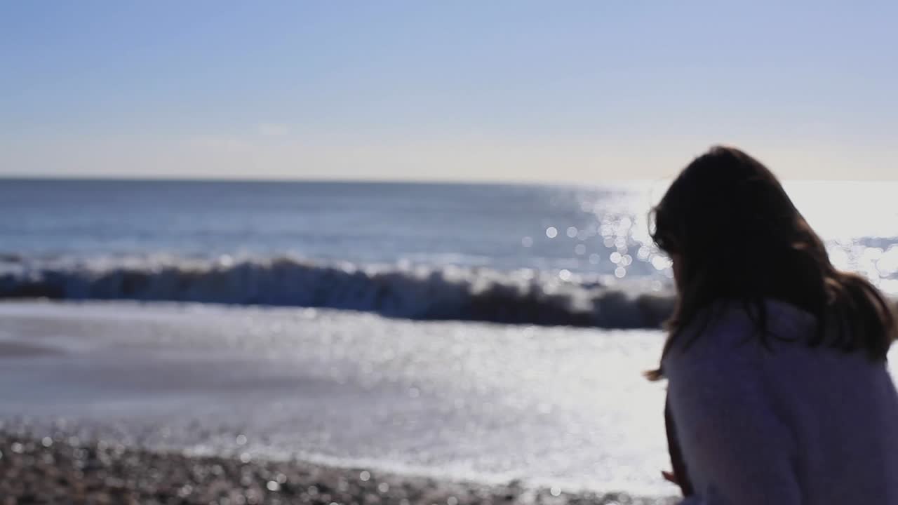 Girl in purple jumper is standing on a sunny beach and picks up a stone, turns around and throws it in a sea. Sun is glaring in camera. Water reflects light. It feels warm. Happy, joyful mood.HD