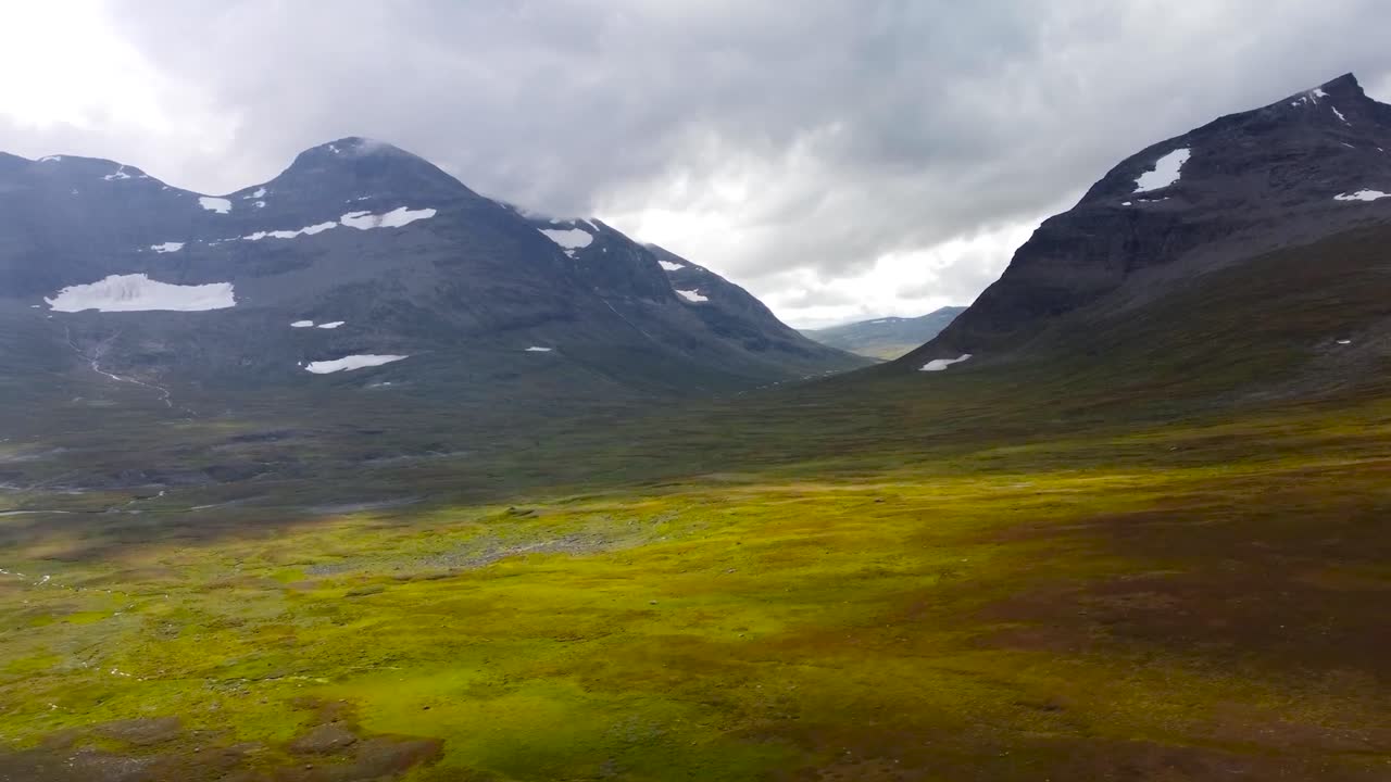 Aerial drone footage slowly flying over golden grassy Sweden landscapes in nature while large dark colored mountains and hills are visible on the horizon and in the back, snow patches on the mountains