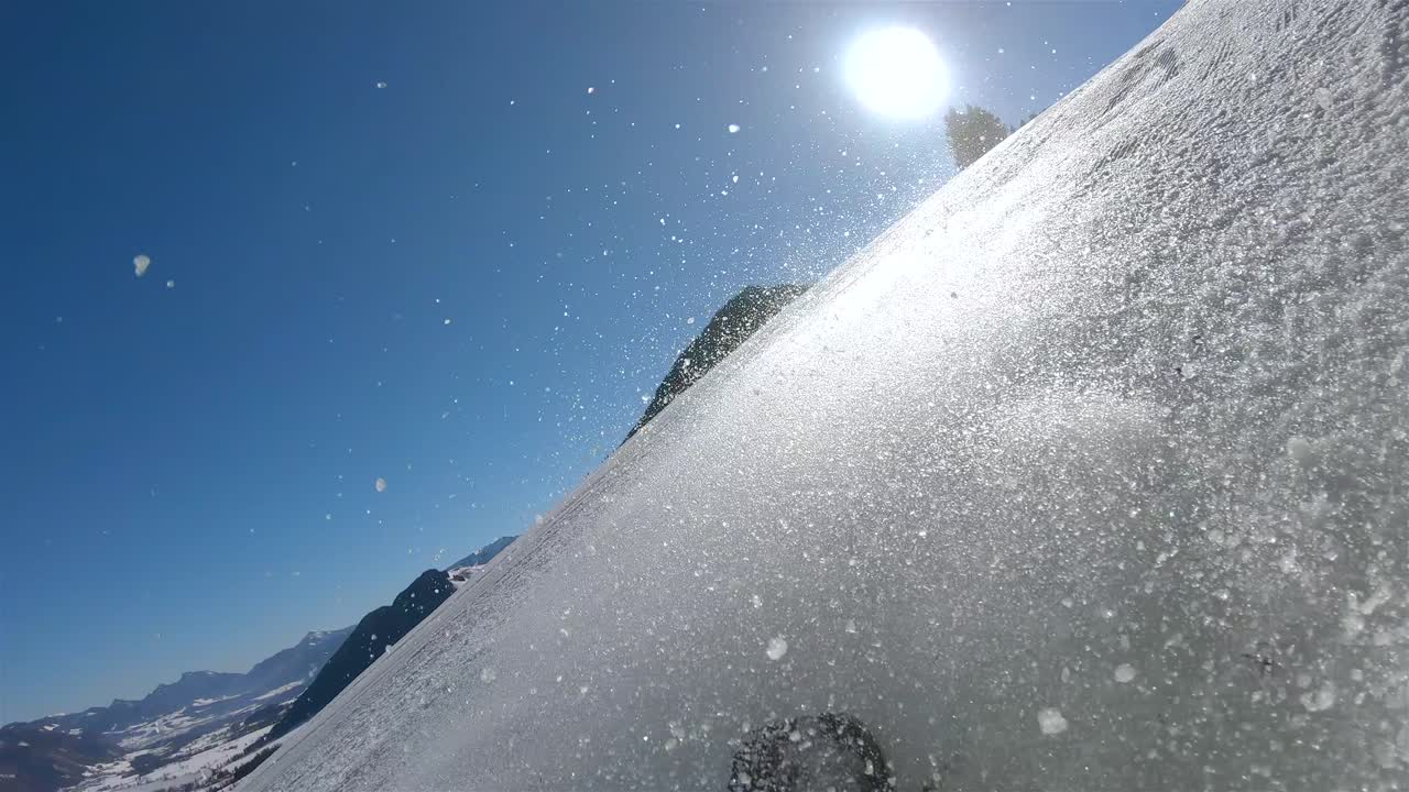 가파른 슬로프를 내려가는 스키어, 배경에는 밝은 태양과 눈이 스키어 뒤에서 솟아오르는 푸른 하늘이 있습니다.