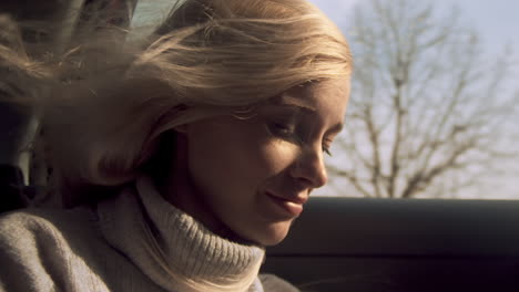 young woman smiles as wind blows long blond hair in car window, close-up