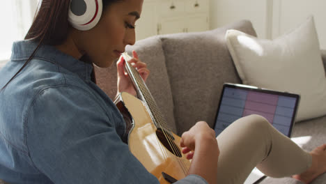 Video-of-happy-biracial-woman-in-headphones-sitting-on-sofa-and-playing-guitar