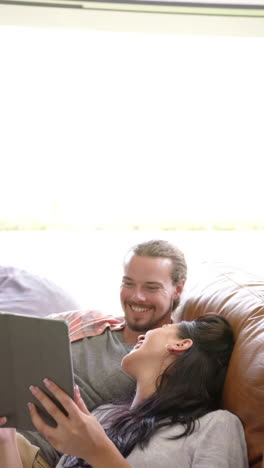 vertical video: laughing couple relaxing on couch, using tablet together at home