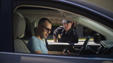 irritated driver with smartphone speaking with police officer zoom in view of angry man finishing phone call then talking and showing driver license to serious woman in police uniform while sitting in car on city street