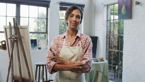 retrato de una artista biracial en delantal sosteniendo pinceles en el estudio, cámara lenta