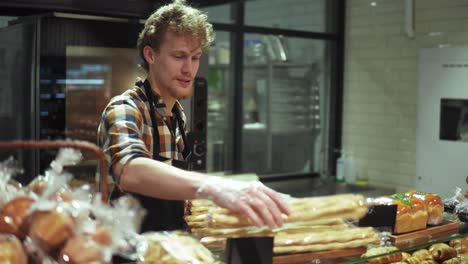 Positive-shop-clerk-in-black-apron-putting-baked-sticks-on-the-showcase-standing-in-the-beautiful-store-with-bakery-products.-Slow-motion