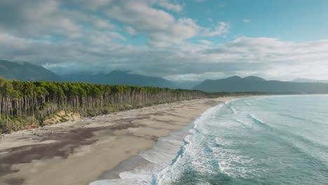 Aerial-drone-view-of-scenic-windswept-Bruce-Bay-with-long-stretching-sandy-beach,-lined-by-native-Rimu-trees-and-rolling-waves-from-the-Tasman-Sea-in-South-Westland,-New-Zealand-Aotearoa