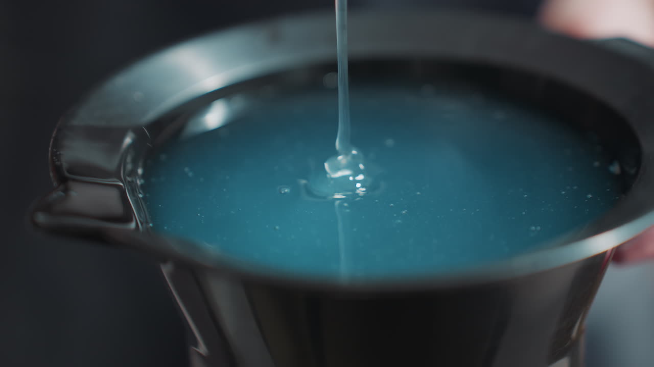 검은색 볼에 푸른색 젤이 쏟아지는 모습이에요. 진한 푸른색 용액이 잔물결을 만들고, 미용실 혼합 볼이 카운터 위에 놓여 있어요. 천천히 부어 윤기 있는 표면을 형성하고, 점성 있는 색소의 클로즈업, 시원하고 분위기 있는 조명이에요.