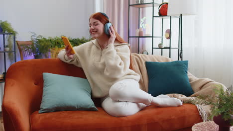 joven feliz con auriculares inalámbricos relajándose sentada en el sofá en casa escuchando su música favorita