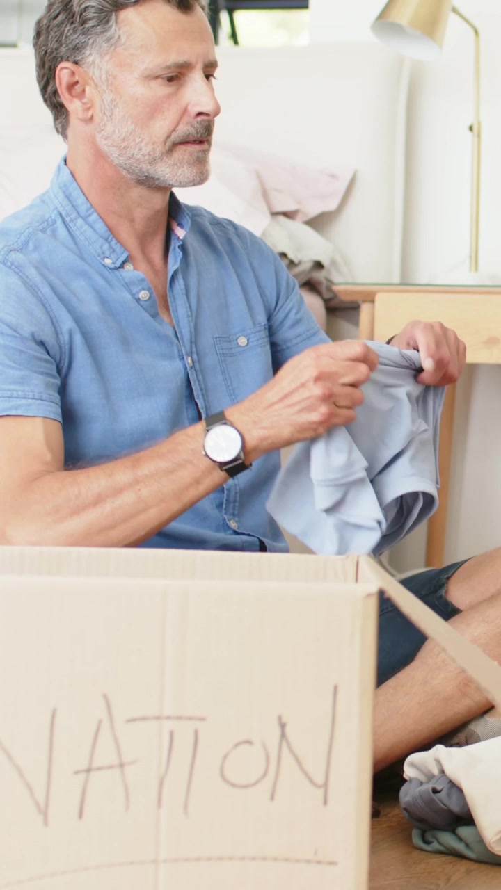 Vertical video: Holding tablet, man offering it, folding light-blue shirt into NATION box at home