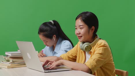 close up of asian woman students wearing headphones and typing on a laptop while sitting with her friend studying on a table in the green screen background classroom