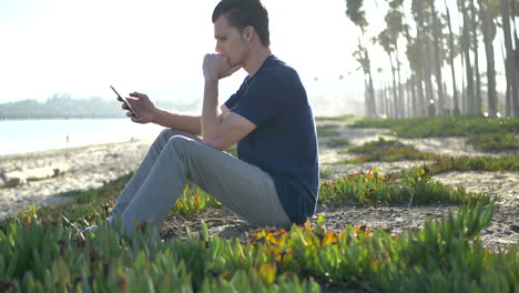 un joven blanco se sienta en la playa mirando su teléfono inteligente mientras parece nervioso, molesto o estresado en santa bárbara, california.