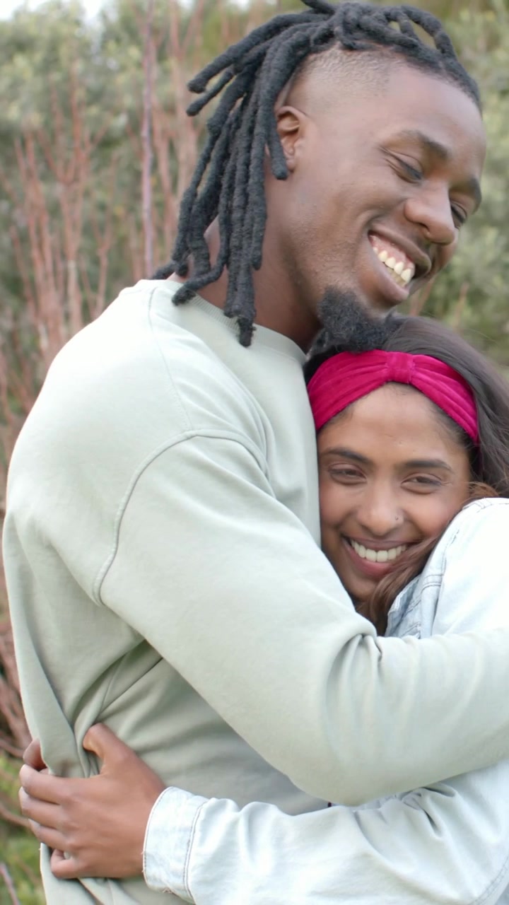 Vertical video: Entering, man lifting brown scarf, draping woman in denim in woods hugging warmly