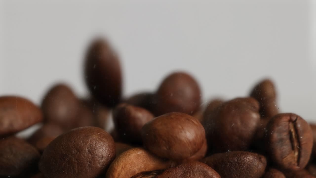 Primo piano di chicchi di caffè che cadono, ripresi al rallentatore con illuminazione soffusa e uno sfondo neutro, che enfatizza la texture e il movimento.