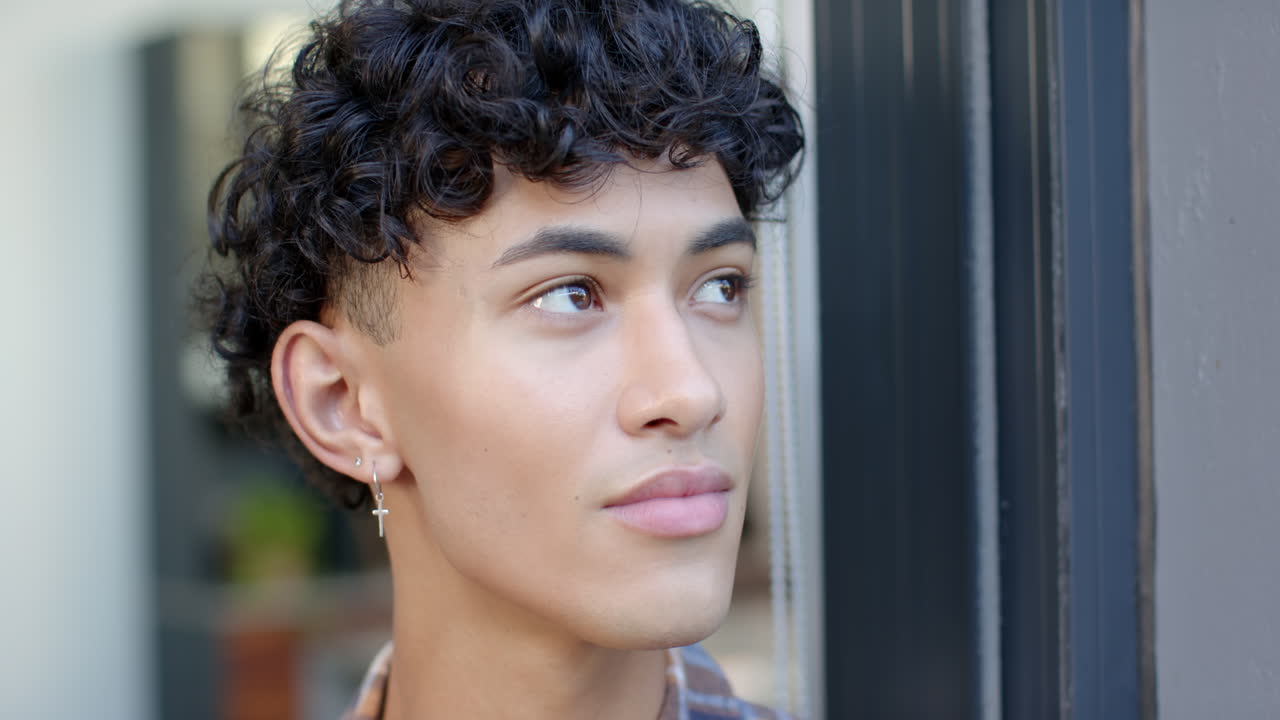 Looking thoughtful, young man with curly hair standing by window at home