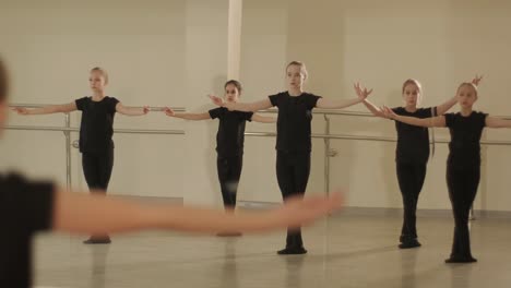 a group of young ballet students in black dancewear practicing positions in a spacious ballet studio with wooden flooring and wall-mounted barres. focused expressions and synchronized movements.