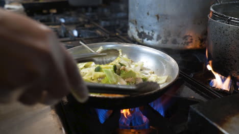 chef uses tongs to stir fry fresh vegetables over flames on commercial restaurant kitchen stove, slow motion hd