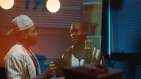 two young stylish guys singers or rappers standing at the mic in the sound studio over the glass window and talking during a record