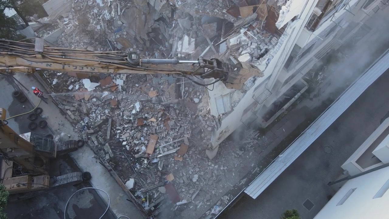 Aerial perspective of active demolition and debris removal in city neighborhood, drone above crane cracking and spinning to remove wall