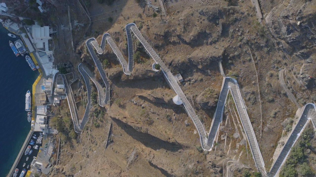 A top-down drone view reveals the famous zigzag switchback path carved into Santorini's caldera cliff, connecting the port to Fira above.