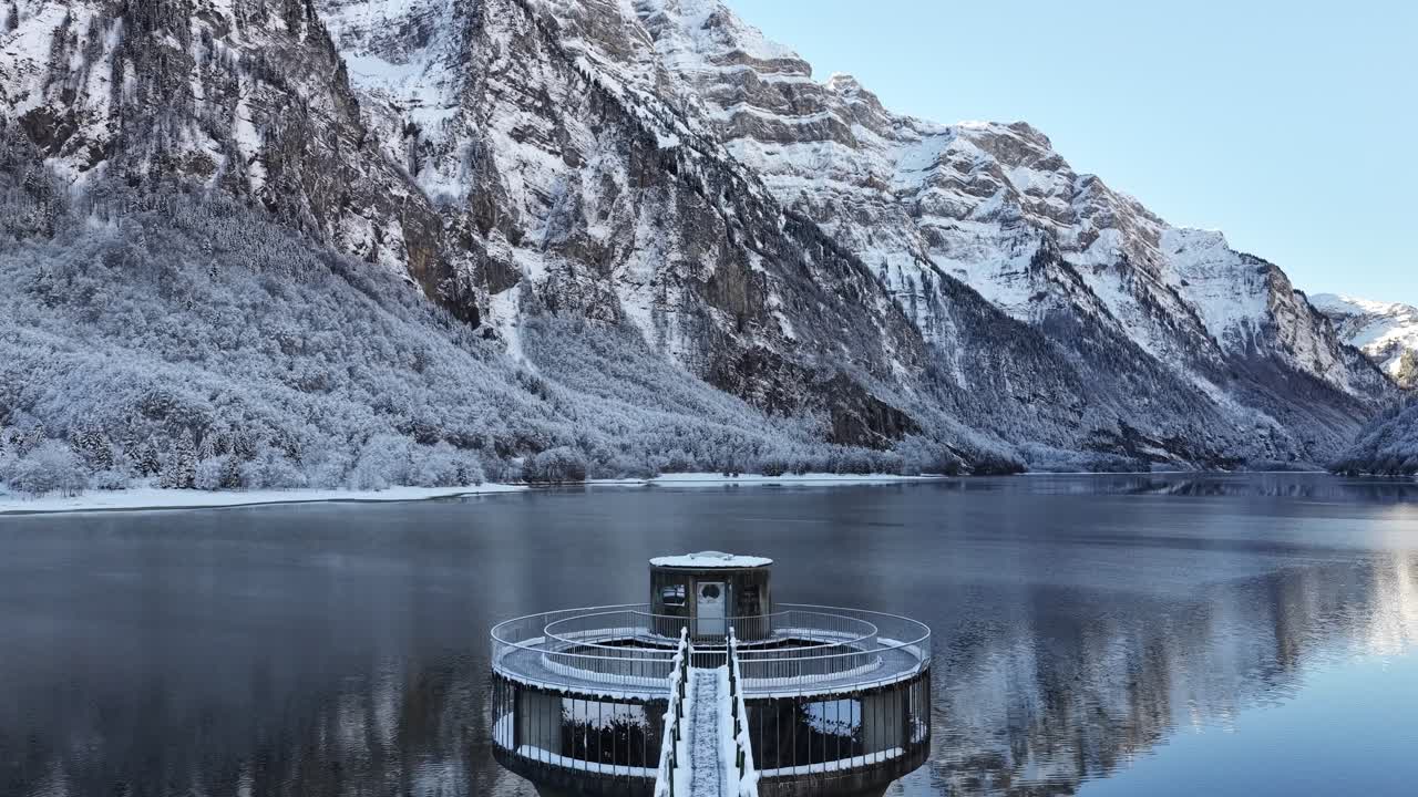 Snow-covered dock extending into a calm alpine lake with snowy mountains reflected in the clear winter water