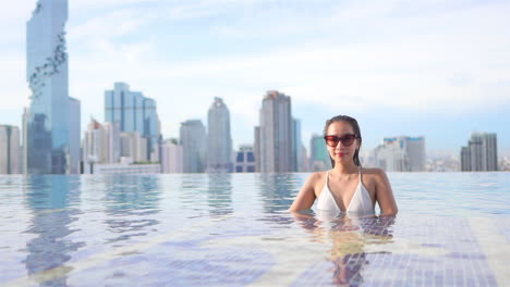 Una-Mujer-Joven,-Respaldada-Por-Un-Horizonte-Urbano,-Se-Relaja-En-Una-Piscina-Infinita-En-La-Azotea