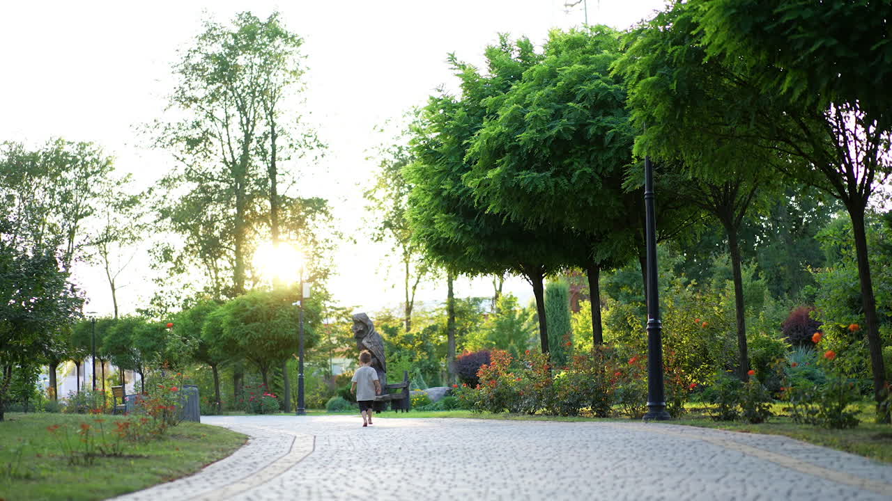 Unrecognized barefoot toddler boy runs by the paved alley in the park. Kid stops far from camera and turns around.