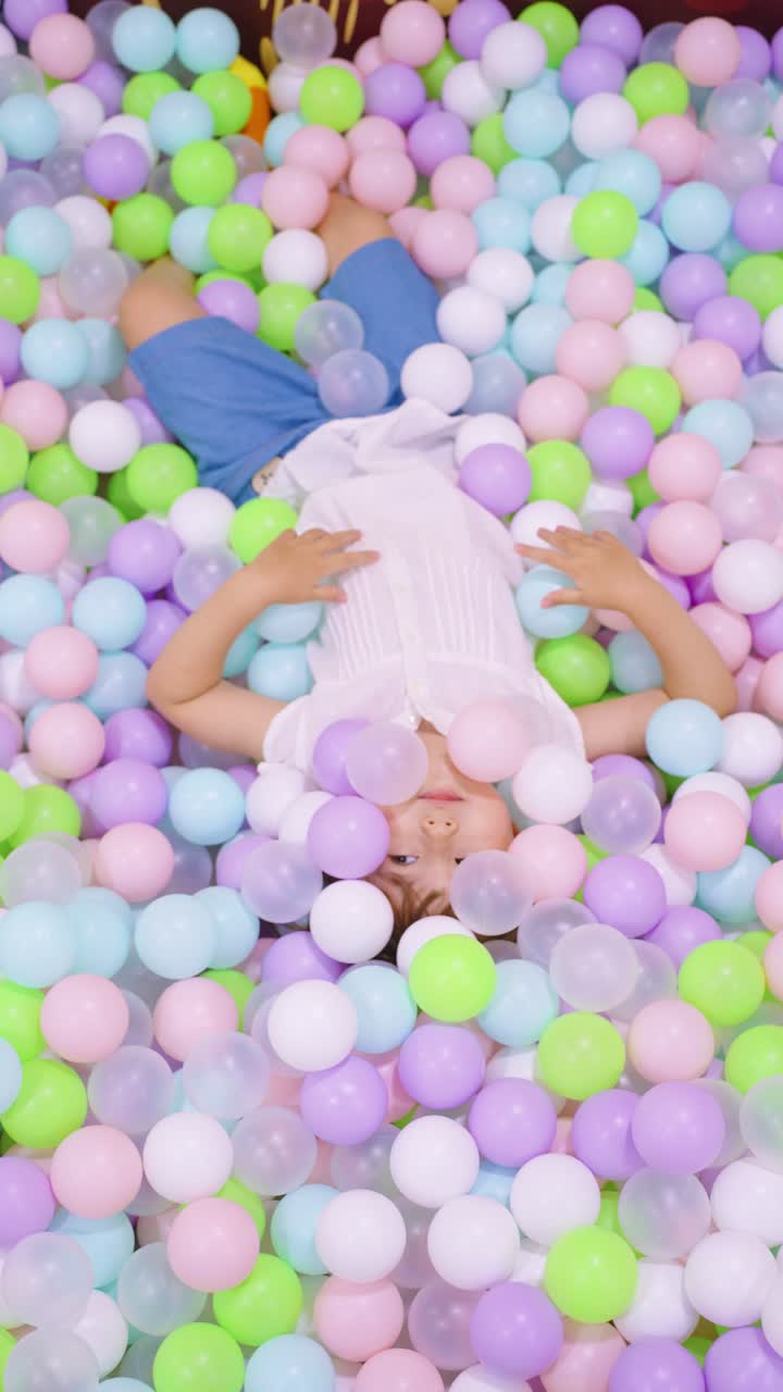 A 5 year old girl lies in a pastel ball pit at an indoor playground, smiling and playing among colorful plastic balls
