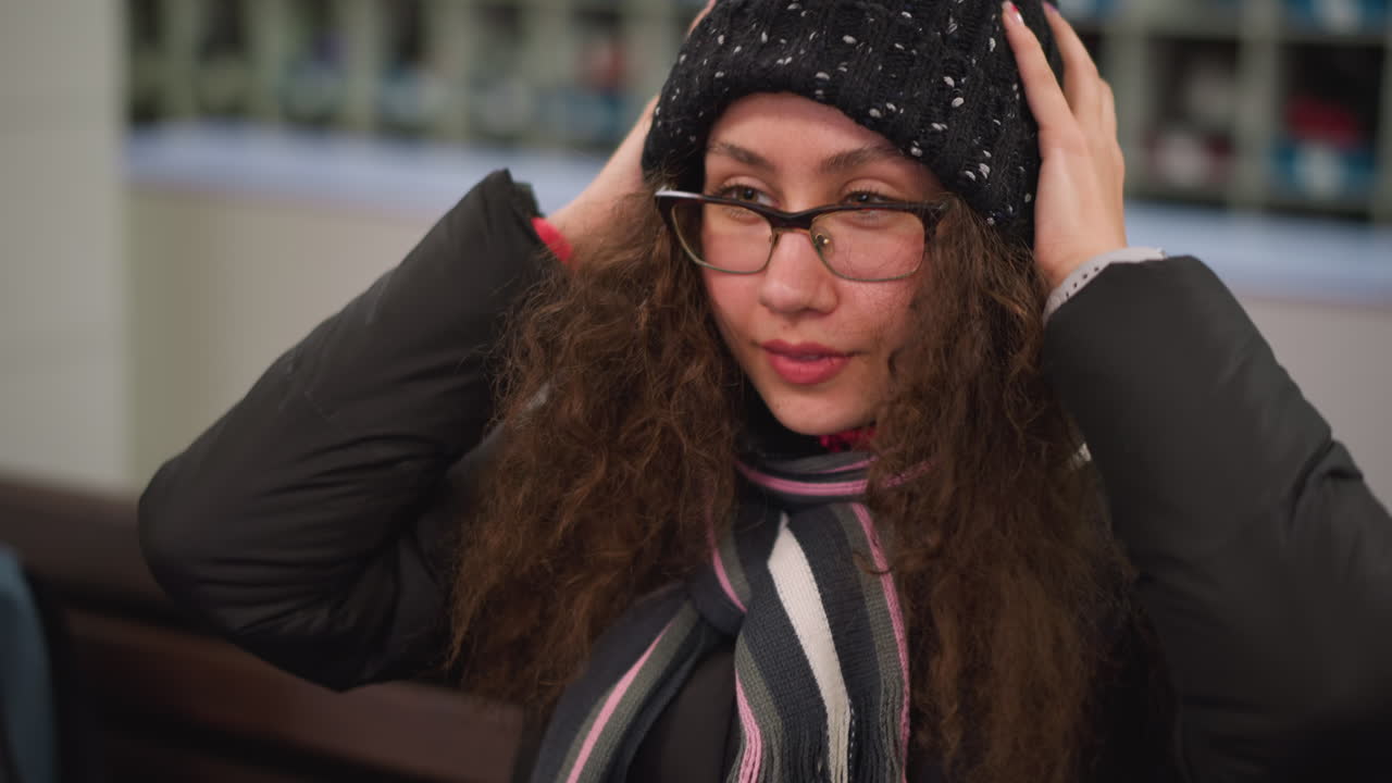 Damsel with long curly hair in black winter jacket and striped scarf bends head forward adjusting hairstyle with both hands, wearing smartwatch, candid authentic indoor moment showing natural beauty