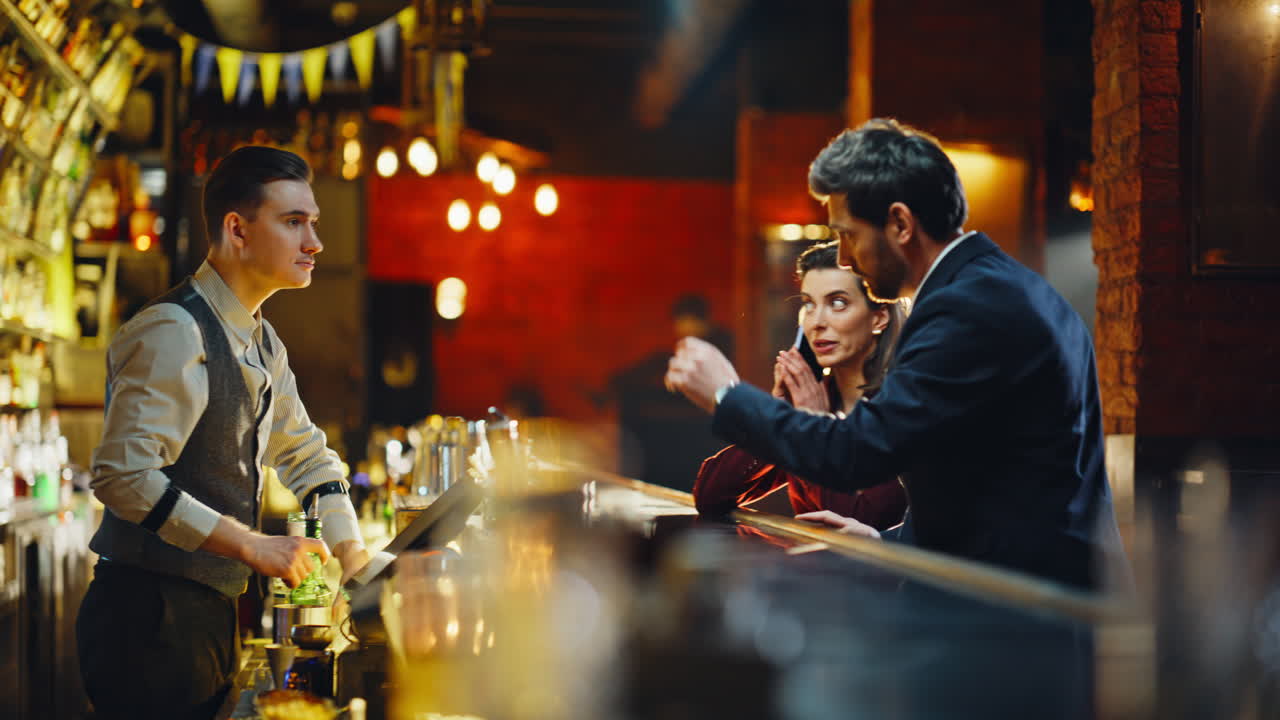 Casal entrando em um bar à noite. Homem conversando com o bartender e pedindo bebidas no balcão.