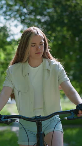 una mujer joven con guantes de motociclista se sienta en una bicicleta al aire libre, mirando pensativamente sus manos mientras está rodeada de exuberante vegetación, el fondo presenta hojas y árboles revoloteantes