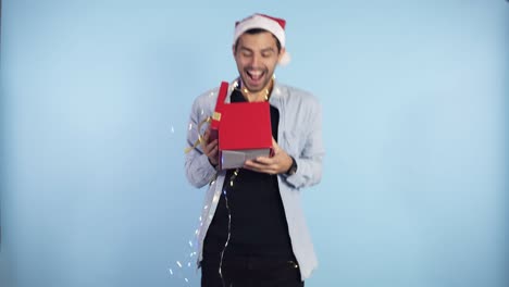 Positive,-young-guy-in-Christmas-hat-and-garland-on-neck-recieves-surprise-from-from-the-side-female-hands---being-in-good-festive-mood,-isolated-over-blue-background.-People,-surprisment-and-presents-concept