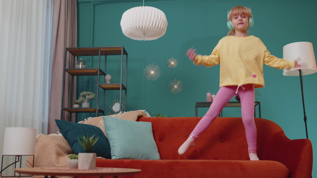 Happy child girl kid in headphones dancing and jumping while listening to the music at home alone