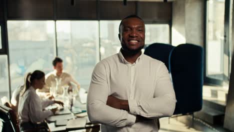 A-successful-businessman-with-Black-skin-in-a-white-shirt-folded-his-arms-on-his-chest,-smiles-and-looks-at-the-camera-against-the-backdrop-of-an-office-team-at-work