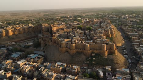 aerial drone shot flying above jaisalmer fort as the sun dips behind the dunes in the distance.