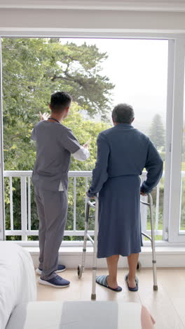 Vertical video of diverse male doctor and senior man using walking frame at home, slow motion