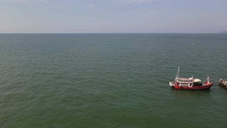 aerial footage sliding towards the left revealing a fishing boat docked with a thai flag flying at the pattaya fishing dock while a boat also speeding on the right, pattaya, thailand