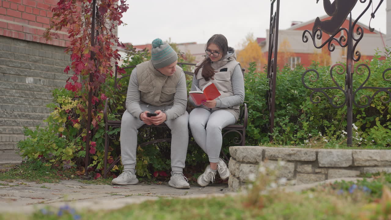 Man using phone seated on bench beside friend reading novel in garden surrounded by autumn leaves and greenery, as he glances at her while she focuses on book during calm outdoor moment