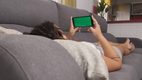 woman on the living room couch watching something on her cell phone, green screen on smart phone