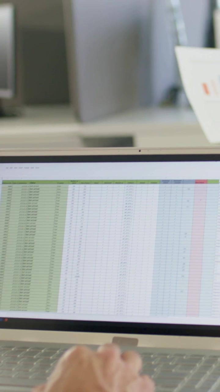 Vertical video: Typing dark-cuff hand on laptop keys checking sheet at cubicle using paper chart