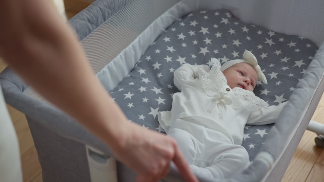 Primo piano di una neonata che dorme pacificamente in una culla con lenzuola a fantasia stellata, la mano del genitore sistema la culla.