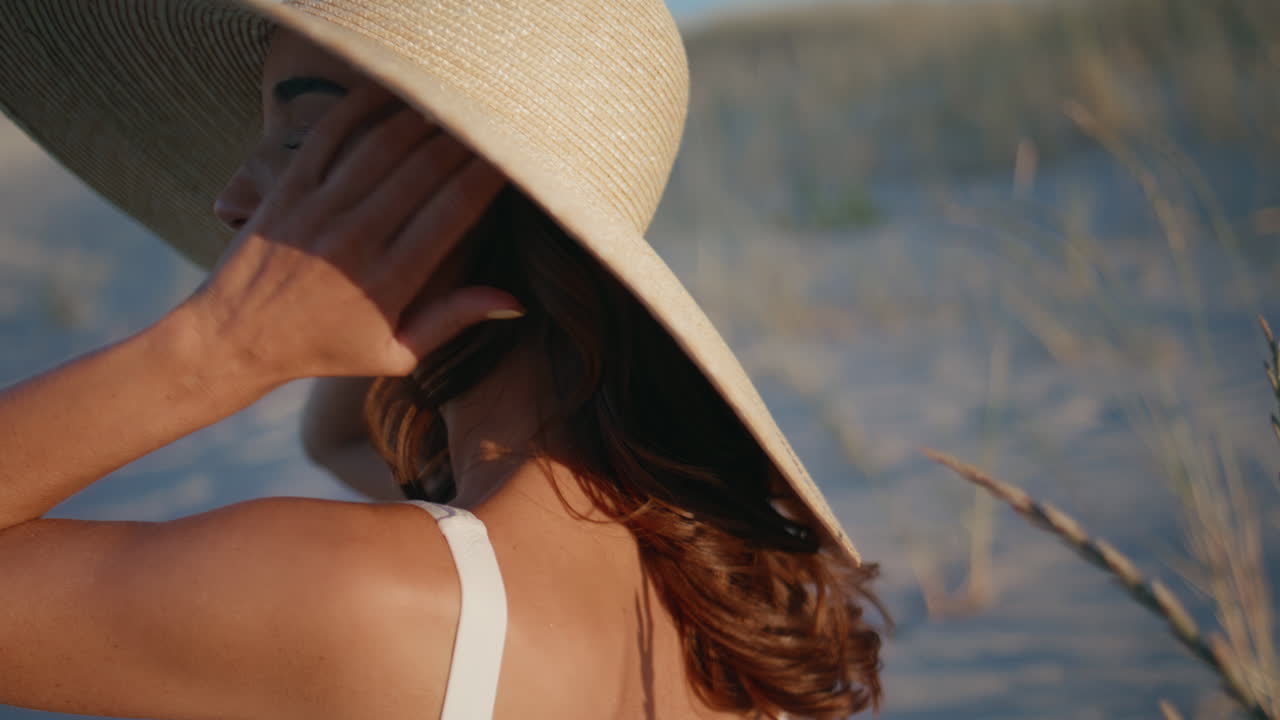 Donna seduta di spalle sulla spiaggia che indossa un costume da bagno, primo piano. Brunetta rilassata con cappello di paglia