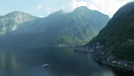 Drone-glides-backward-over-the-stunning-lake-revealing-Hallstatt-village,-nestled-amidst-colossal-mountains-that-throw-it-into-the-afternoon-shadow,-as-cruise-boats-approach-the-docks