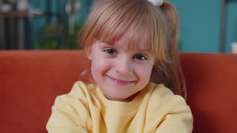 smiling child girl kid on home sofa looking at camera makes heart gesture demonstrates love sign