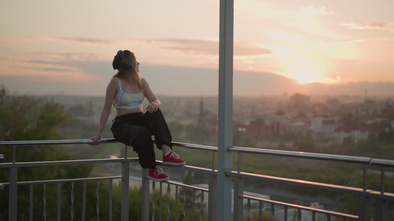 Ballerina al tramonto in una città vivace. Ballerina classica in scarpe da ginnastica interagisce con il tramonto sullo sfondo di un vivace paesaggio urbano. Artista festeggia il crepuscolo salutando con le cuffie tra i grattacieli colorati